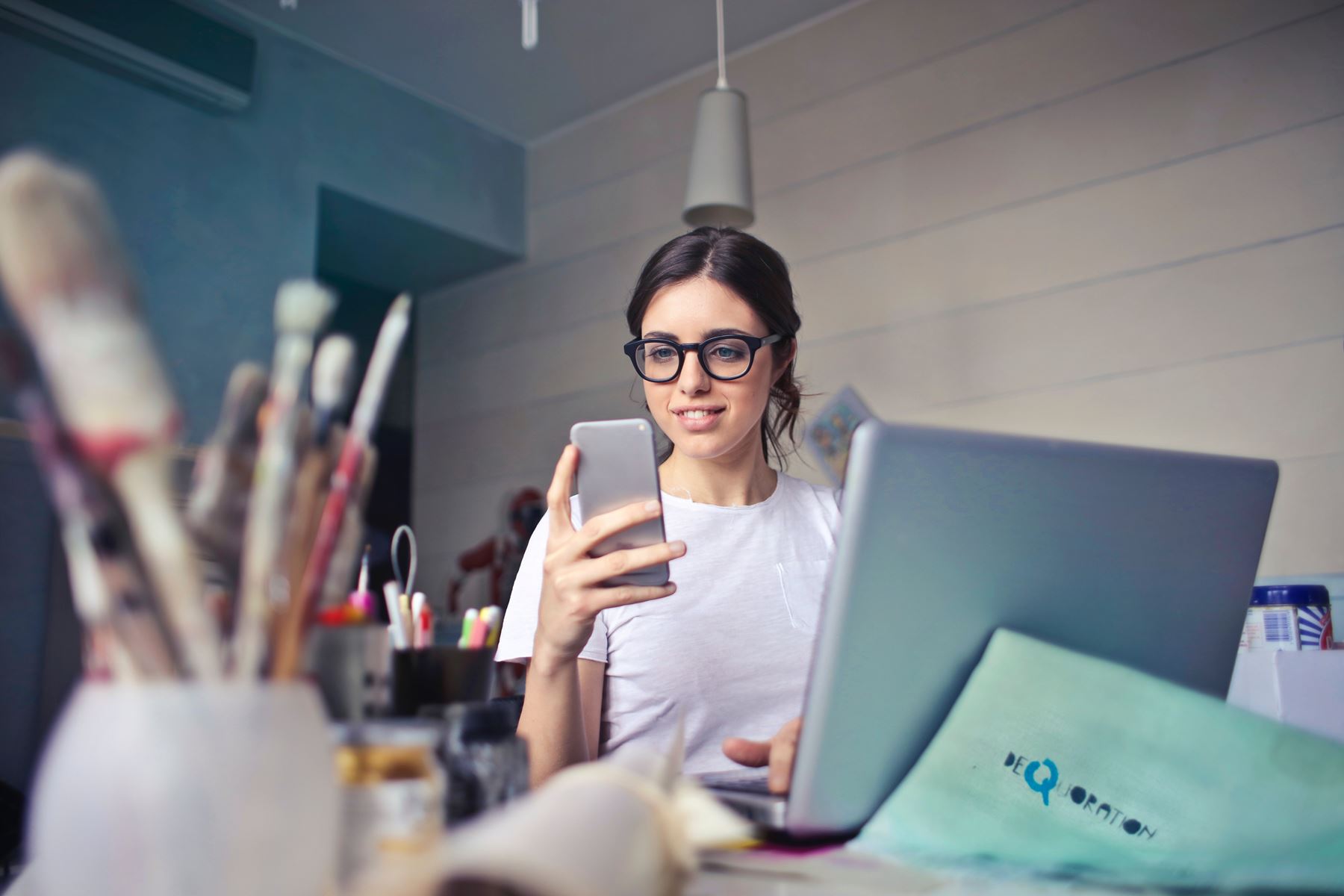 Girl holding and looking at her cell phone while one hand is on the keyboard of her laptop