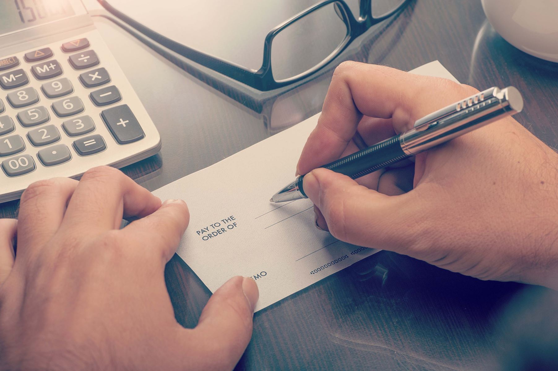 Hands, one hand is holding a check and the other is writing out the check. There are glasses and a calculator on the table