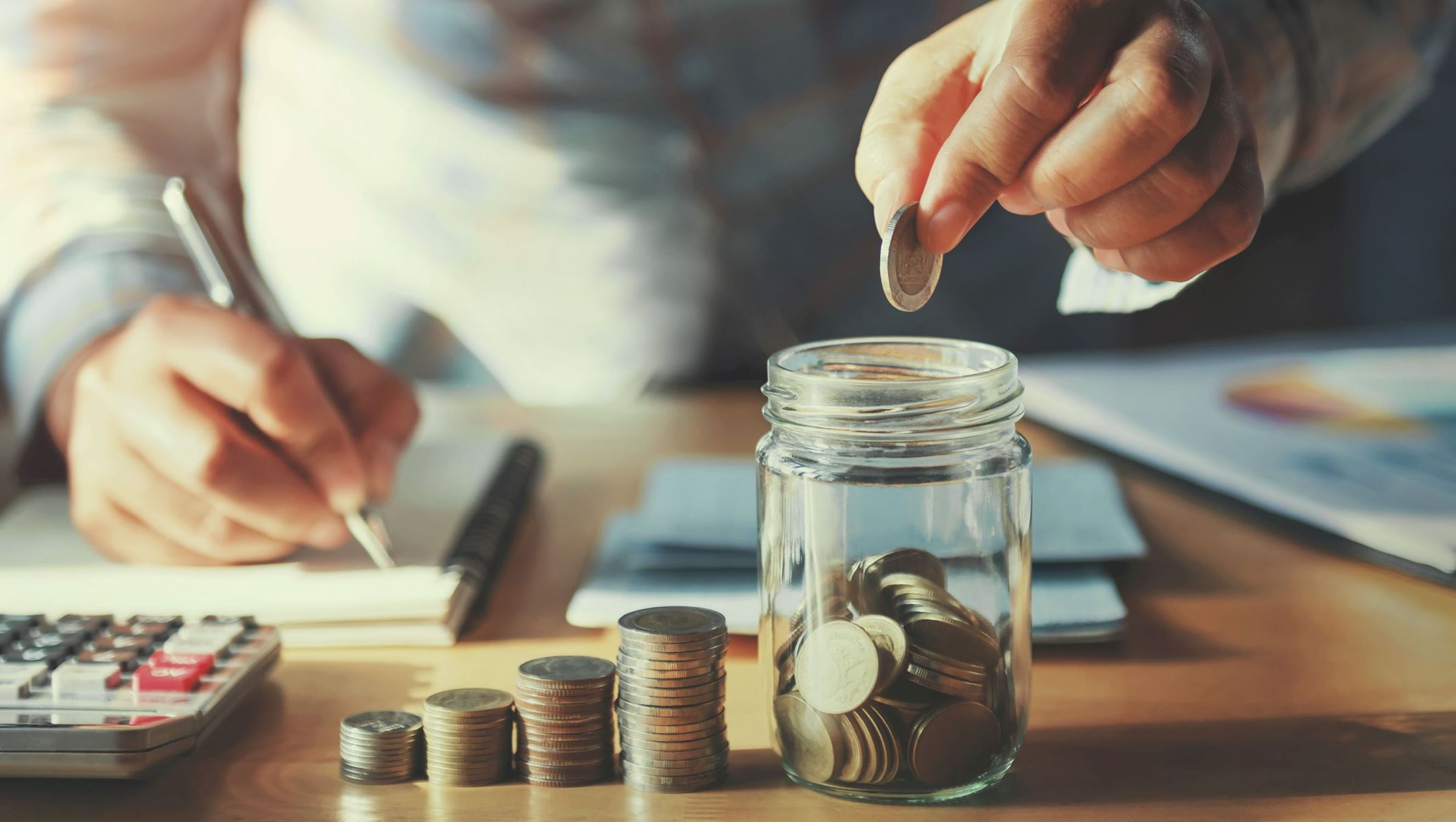 Coins stacked in a graph type shape, there is a glass jar and a man is putting a coin in it. He is also writing on a notepad